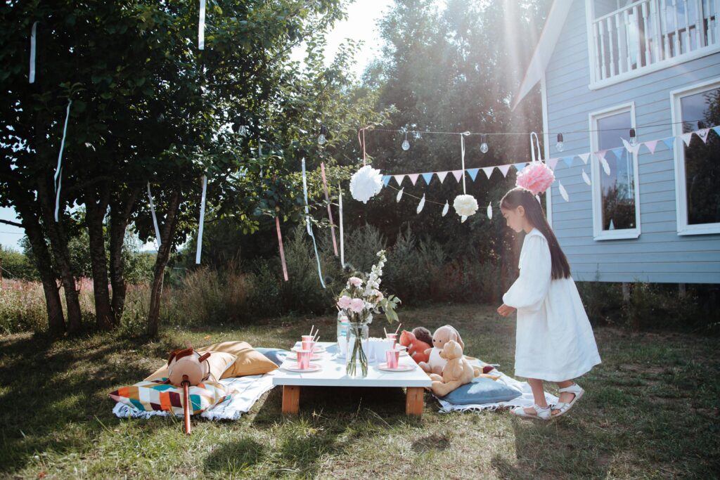 Mädchen am geschmückten Kindertisch im Garten wo schon ihre Kuscheltiere sitzen. Alles ist bunt aber nicht überladen als Deko zur Einschulung oder zum Geburtstag.