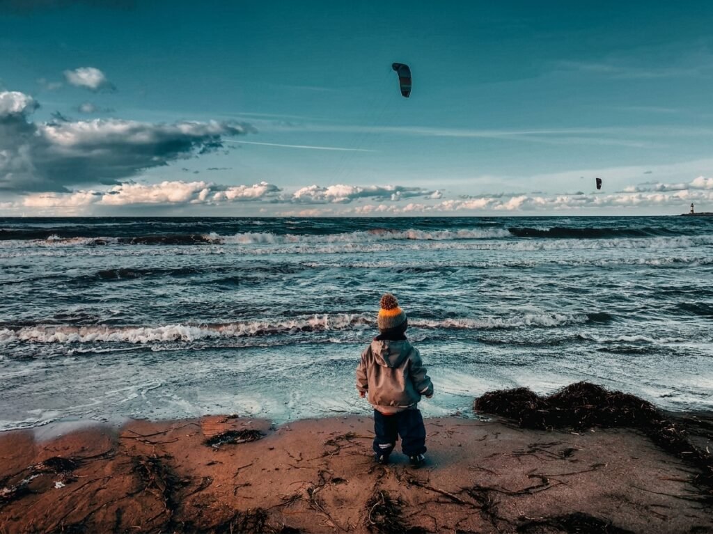 Kind steht warm angezogen vor rauer Ostsee mit Kitesurfern